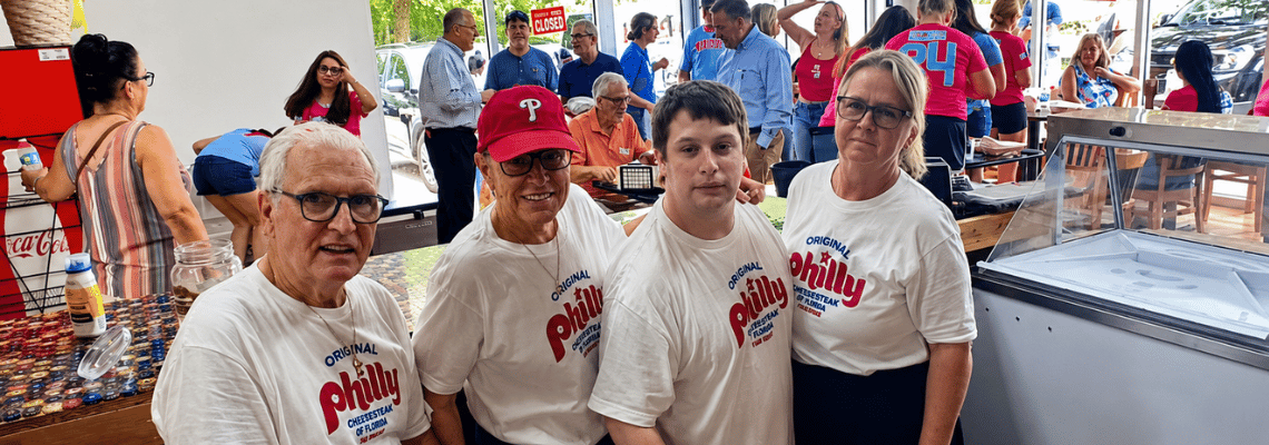 Bryan Keenan, owner Mary Keenan, Dustin Concordin, and Helen Szymczak pose together inside Original Philly Cheesesteak of Florida during the restaurant’s grand opening celebration in Palm Coast as customers, local supporters, and community leaders gather throughout the dining area in the background.